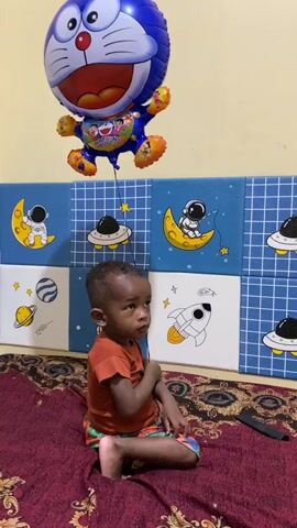 A young boy sits on a red patterned blanket. He is wearing an orange t-shirt and colorful shorts. He is holding a string that is attached to a Doraemon balloon, which floats above him. The background features a wall with blue and white square panels decorated with space-themed images, including astronauts, moons, planets, and UFOs. The boy appears to be looking up at the balloon.