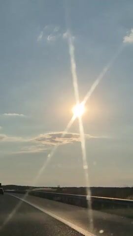 This video shows a view from a moving vehicle on a highway. The sun is bright and high in the sky, creating lens flares that streak across the frame. The sky is a clear blue with scattered white clouds. On the right side of the road, there's a guardrail and a field with some sparse vegetation. In the distance, a line of trees can be seen. The road itself is asphalt with clear white lane markings. A single car is visible in the distance, also on the highway. The overall impression is of a bright, sunny day, likely late morning or early afternoon, experienced while traveling on a road through a rural or semi-rural landscape. There is no visible text in the video.