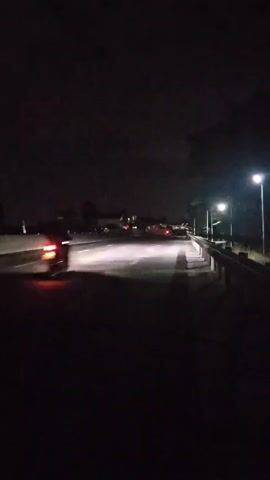 The video depicts a nighttime scene of a road illuminated by streetlights, with a single motorcycle visible in the distance. The road appears to be a bridge or elevated roadway, as indicated by the presence of guardrails along the side. The sky is dark and cloudy. The motorcycle has its headlight on and tail light is visible as it moves along the road. The foreground is dark, with the shadow of the guardrail visible on the road surface. There is a sparse tree visible on the right-hand side of the frame. The scene is quiet, with no audible sounds.