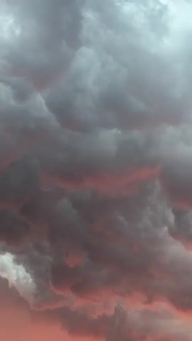 A video shows a sunset with dramatic clouds. The clouds are dark gray and textured, with streaks of pink and orange light filtering through them from below. The bottom of the frame shows the silhouette of rooftops and some trees. The sky is the primary focus of the video.