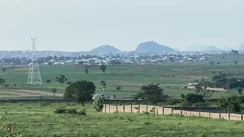 This media file captures a panoramic view of a rural landscape in Gidan Waziri, Nigeria. The scene is characterized by rolling green hills and agricultural fields, dotted with scattered settlements and distinctive rocky outcrops. In the foreground, a low concrete wall with a fence runs horizontally across the frame, separating the viewer from a cultivated field. Various trees and shrubs are interspersed throughout the landscape, adding texture and depth.

In the mid-ground, a small cluster of houses with white roofs are visible. Further into the distance, a larger settlement with numerous buildings and pitched roofs stretches across the horizon, partially obscured by haze. The backdrop is dominated by several large, rounded hills that rise majestically against a pale, overcast sky. A tall transmission tower is also visible in the distance, suggesting the presence of infrastructure within the wider area.

The time of day appears to be daytime, likely late morning or early afternoon, given the diffused light. The weather seems to be hazy or overcast, contributing to the soft, muted tones of the landscape and the distant visibility. No specific activity or event is evident; the scene conveys a sense of quietude and rural life. No visible text is present in the media file.