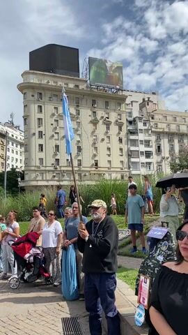 The video shows a daytime scene in a city square with a crowd of people, mostly adults. The group is diverse in appearance, with individuals of various ethnicities, ages, and clothing styles. Some are wearing sunglasses and hats. Several people are clapping and looking toward what appears to be a speaker or event location, which is off-camera. A man in the foreground holds an Argentinian flag. A few people are pushing strollers. The background features large, multi-story buildings in a European style, one of which has a "Sarmiento" billboard on top. The sky is partly cloudy.