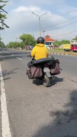 A person wearing a yellow hoodie rides a motorcycle on a paved road. The motorcycle has two saddlebags attached to the rear. The rider is facing away from the camera. The road has a white line painted on the right side and a painted white line with a dashed white line on the left. Trees line the left side of the road, casting shadows on the pavement. In the background, there are more trees, a building with a red roof, power lines, and other vehicles. The sky is blue with scattered clouds. The overall scene appears to be a daytime outdoor shot of a person traveling on a road.