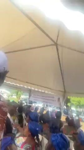 The video shows a large crowd of people sitting under a white tent. The crowd appears to be mostly women. A banner is visible in the background and also some white SUV-type vehicles parked outside the tent. The audio includes voices and general background noise.