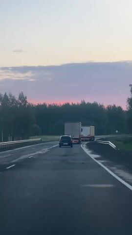 The video shows a view from a vehicle driving on a highway at dusk. In the foreground, the road surface is visible, appearing wet and marked with white lines. In the middle distance, there is a black car driving in the same direction, followed by two large trucks. The trucks are side-by-side, with the truck on the right slightly ahead. The truck on the left has a white trailer, and the truck on the right also appears to have a white trailer. Beyond the trucks, there is a dense line of trees forming a forest. The sky above is a pale pink and purple, indicating either sunrise or sunset, with some clouds. There are guardrails visible on the sides of the road, and a signpost with a circular red and white symbol on the left side of the road. The overall atmosphere is peaceful and serene, typical of a drive during twilight.