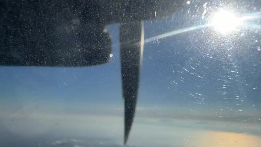 The video shows a close-up of a rotating propeller blade seen through a dirty airplane window. The sun is visible in the upper right corner of the frame, creating lens flare and reflecting off the window. The propeller blade is dark and blurred due to its rapid rotation, with a lighter, reflective section catching the sunlight. Below the propeller, a hazy blue sky and white clouds are visible, indicating the plane is in flight at a considerable altitude. The window itself is marked with numerous scratches and smudges, obscuring the view of the outside world. The overall impression is one of motion and light filtering through a weathered surface.