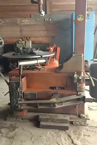 A person is working in what appears to be a garage or workshop. The main object of focus is a large, orange and red industrial machine, likely a tire-changing machine, with various hoses, gauges, and a large black tank attached. In the background, there are wooden walls, shelves, and other equipment. A person, wearing beige pants and a dark shirt, is seen interacting with a car tire, moving it around the machine. The floor is concrete, and the overall environment suggests a functional, working space.