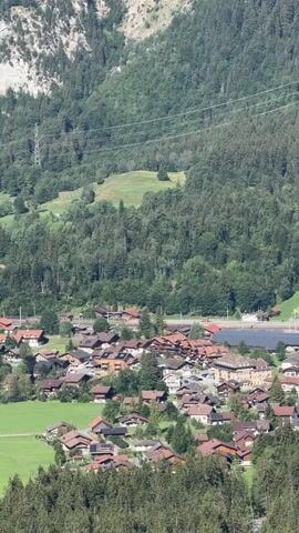 The video is a vertical panoramic shot of a Swiss village nestled in a valley. The village consists of traditional chalets with red roofs, surrounded by green fields and dense pine forests. Towering mountains with rocky cliffs and snow-capped peaks form the backdrop. The sky is clear blue with scattered white clouds. The video begins with a close-up view of the village and gradually zooms out to reveal the expansive mountainous landscape. There are no people or animals visible in the video. The overall impression is one of natural beauty and tranquility.