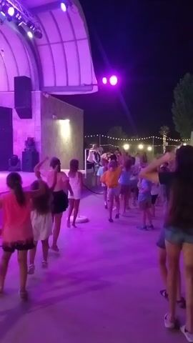 A group of children and young people are gathered outdoors at night, illuminated by purple and pink stage lights and string lights. A stage with a band and performers is visible on the left, with a white canopy overhead. The audience is lined up in front of the stage, with some people dancing and others watching. The ground appears to be paved.