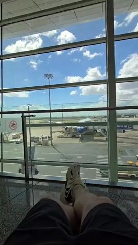The video shows a first-person view of a person relaxing in an airport waiting area, with their legs crossed and extended towards a large window. Through the window, a busy airport tarmac is visible under a bright, cloudy blue sky. Several airplanes are parked at gates, with jet bridges attached. Various airport vehicles, including baggage carts and what appear to be taxis, are present on the tarmac. The interior of the waiting area features a polished floor and modern architectural elements, including large glass panels and metal structural beams. The overall scene conveys a sense of travel anticipation or waiting for a flight.