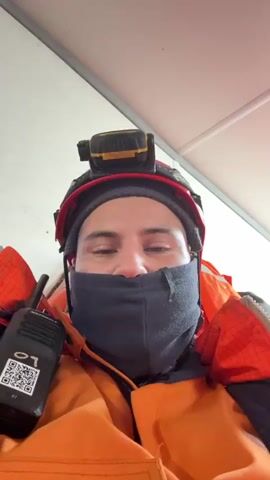 The video begins with a close-up of a person wearing a red helmet with a headlamp, an orange life vest, and a black face mask. The camera moves, revealing a room with a man in an orange jumpsuit and reflective jacket standing and talking. The room has a white ceiling with fluorescent lights and a security camera. Behind the man are lockers and other equipment.