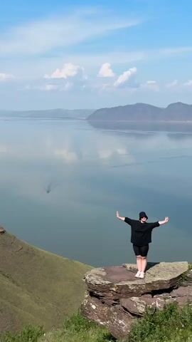 A person stands on a rocky outcrop overlooking a large body of water. The person is wearing a black t-shirt, black shorts, a black hat, and white sneakers. They have their arms outstretched to the sides, palms facing forward, and their head tilted slightly upwards. The person is smiling. The body of water is calm and reflects the sky. In the distance, mountains are visible, and a small boat is leaving a wake on the water. The sky is blue with scattered white clouds. The terrain in the foreground is green and grassy with some rocks. The camera is positioned at eye level with the person and is looking slightly down towards the water. The video pans slowly to the right, showing more of the landscape.