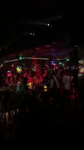 The video shows a dimly lit, crowded nightclub. People are standing and sitting at tables, some are talking, and others appear to be dancing. There are bar stools and tables set up, with drinks and bottles visible. The environment is dark with colored lights, including red, green, and blue spotlights, illuminating the space and creating a party atmosphere. There are also neon signs visible in the background. Some individuals are smoking. The overall impression is of a lively social gathering in a club setting.