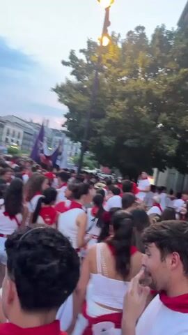 A large crowd of people wearing white shirts and red bandanas are gathered in a city square. The crowd is moving and some people are holding up phones to take pictures. There are buildings in the background and trees. The sky is cloudy.