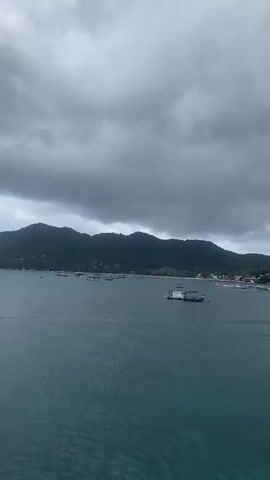 The video opens with a wide shot of a bay under a cloudy sky, revealing a mountainous island covered in vegetation in the background. Several boats dot the water, with a cluster appearing closer to the shore, indicating a small town or village. The camera then moves forward, closer to the island, focusing on the village as it comes into view. Finally, the camera pans up to show the mast of the boat on which the video is being recorded, revealing a Garmin radar system and other communication equipment. Two individuals wearing red life jackets are seen briefly near the bow.