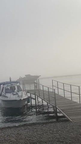 A video shows a foggy scene at a body of water. In the foreground, a wooden pier extends into the water. On the left, a white boat is docked. Further out on the water, a larger boat is moving. A person is walking on the shore near the pier, and another person stands on the pier. The overall atmosphere is hazy and subdued due to the fog.