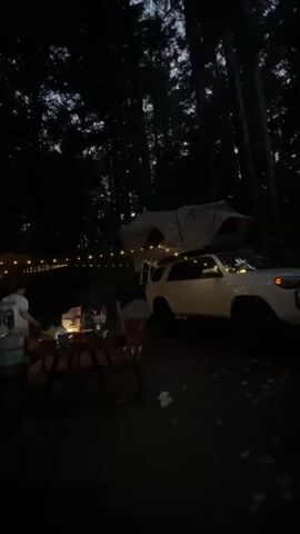This video captures a campsite at dusk. In the foreground, a picnic table is set up with several people gathered around it, some appearing to be preparing food or drinks. String lights are visible, stretched between trees, providing illumination. A white SUV with a rooftop tent is parked to the right of the picnic area, and another white SUV is visible to the left. The scene is set within a dense forest, with tall trees forming a dark backdrop against the twilight sky. The overall atmosphere is relaxed and communal, suggesting a camping trip or outdoor gathering.