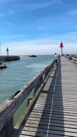 The video shows a wooden pier extending into a body of water under a clear blue sky with a few white clouds. On the pier, a person wearing a white shirt and dark pants stands near the railing, looking out at the water. Further down the pier, another person, wearing a white shirt, is also visible. In the water, a red sailboat with the number 611 on its side is sailing. There are two people on the sailboat. The sailboat is moving from left to right in the frame, leaving a wake behind it. On the left side of the frame, a wooden structure, possibly a breakwater or pier, extends into the water, and a green lighthouse is visible at its end. Further out in the water, a white lighthouse with a red top is also visible. A small red boat with a person in it is moving towards the breakwater on the left. The water appears calm with gentle ripples. The overall scene depicts a sunny day at a coastal location with maritime activity.