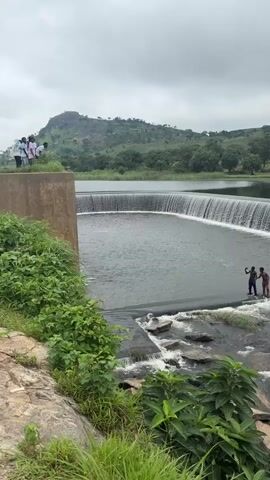 The video shows a large man-made dam with water cascading over its edge. On the left side of the dam, a group of people are standing on a rocky ledge, looking towards the water. On the right side of the dam, two young males are standing on the edge of the dam, near where the water flows over. The background consists of lush green vegetation and rolling hills under a cloudy sky. The water is a dark gray color.