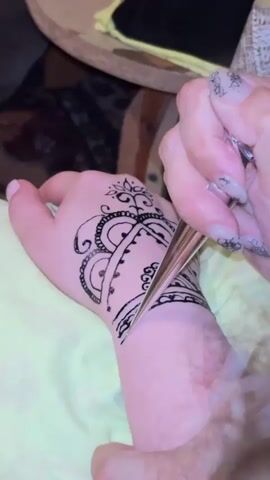 A close-up shot shows a hand with a henna tattoo being applied to the back of the wrist and hand. The henna paste is being applied from a cone-shaped applicator by a person whose hand is partially visible holding the cone. The tattoo design is intricate, featuring a central floral motif with swirling patterns and decorative lines. The hand receiving the tattoo is resting on a yellow fabric. The background is blurred, showing hints of wooden furniture and fabric.