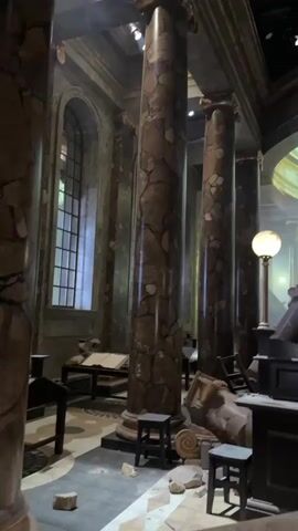 The video shows a dimly lit, grand interior space with large marble columns and an arched ceiling with a large window. In the foreground, there is debris scattered on the floor, including what appears to be broken stone and furniture. A large, ornate desk or counter is visible, with a dark-colored mechanism on top of it, possibly part of a display or prop. Several small stools are placed around the area. Smoke or fog drifts through the scene, obscuring parts of the background. Two large, crystal chandeliers hang from the ceiling. The overall impression is that of a damaged or ruined grand hall, possibly a set from a film or theatrical production, given the staged appearance of the destruction and lighting. No people are visible in the footage.