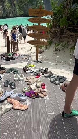 The video opens with a wide shot of a tropical beach. Numerous pairs of sandals and flip-flops are neatly arranged on a wooden boardwalk that leads to the sand. In the background, people are visible walking on the beach and swimming in the turquoise water. A wooden signpost with multiple directional arrows is also present. The camera then pans and zooms to focus on a woman wearing a black and white patterned robe walking past the shoes. She briefly stops and touches the signpost. The shot then shifts to show another woman, wearing a wide-brimmed hat and a light green two-piece outfit, adjusting her hat. Other people are seen walking towards and away from the beach. The camera continues to show the collection of footwear and glimpses of other tourists enjoying the beach environment. The overall scene depicts a popular tourist destination where visitors are required to remove their footwear before accessing certain areas.