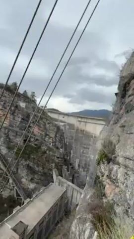The video shows a point-of-view shot of a person engaged in a climbing activity near a large concrete dam structure in a rocky, mountainous area. 

The scene starts with a wide shot of the dam structure, then the camera angle shifts upward to focus on the rocky cliff face. Climbing ropes are visible, anchored to the rock face with metal fixtures. The camera seems to be attached to the climber, following their movement as they navigate the rocky terrain.

Vegetation, including small bushes and trees, grows on the cliffside. A deep canyon or valley stretches out below, with a river or body of water visible at the bottom. The weather appears cloudy and overcast.

Overall, the video depicts a challenging outdoor climbing activity in a natural setting, with the imposing presence of the dam structure in the background.