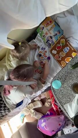 The video shows two young girls playing together in what appears to be a makeshift fort. They are both lying on a white sheet, surrounded by blankets and pillows. On the sheet in front of them is a collection of colorful stickers and a wooden board with pictures of fruits and professions. The girl in the foreground has purple-tipped hair and is wearing a white t-shirt and bracelet. She is reaching out to place a sticker. The girl behind her has blonde hair tied in a ponytail with a yellow clip and is also reaching for stickers. There are several pairs of children's shoes scattered around, as well as a large, sparkly purple toy. The environment appears to be a home, possibly a living room or bedroom. The lighting suggests it is daytime.