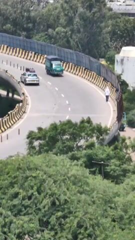 The video shows an aerial view of a road with vehicles and a person. The road curves to the left and is bordered by a yellow and black striped barrier. On the road, a small green truck is moving forward, and a person in a white shirt and dark pants is walking along the right edge of the road. Further ahead and to the left, a white car is visible. In the foreground, lush green trees obscure the lower part of the scene. The overall environment appears to be an outdoor, possibly urban or semi-urban setting, with the road elevated.