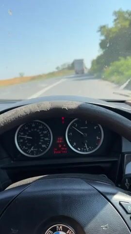 The video is a first-person perspective from inside a car driving on a road. The driver's hands are on the steering wheel, which is worn. The dashboard is visible, showing the speedometer and tachometer. The car is following a truck on a two-lane road with a white line dividing the lanes. The surroundings are rural, with trees and vegetation on the right side of the road and fields on the left. The sky is clear and blue, indicating a sunny day. The video captures the movement of the car and the scenery passing by, with a focus on the road ahead and the dashboard instruments.