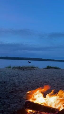 A campfire burns on a sandy beach. Beyond the beach, a calm body of water stretches to a tree-lined horizon. The sky above is a deep blue with scattered clouds. A single person is visible in the water, riding a jet ski. The jet ski is creating a small wake behind it. The scene appears to be set at dusk or dawn.