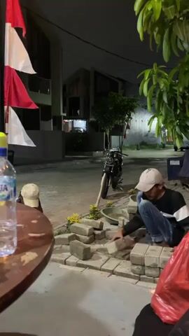 The video is shot at night. In the foreground on the left, a wooden table with a bottle of water and a cap is visible. In the middle ground, two people are seen crouching on the ground. One person, wearing a white cap, blue jeans, and a black and white long-sleeved shirt, is placing bricks. The other person, wearing a beige cap, is partially visible on the left. A motorcycle is parked in the background, and flags are displayed on the left side of the scene. The environment appears to be an outdoor area with buildings and trees.