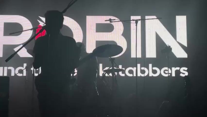 A band performs on stage in front of a large illuminated screen. The screen displays the word "ROBIN" in large white letters, with "and the Ketabbers" in smaller white letters below. A silhouette of a guitarist is on the left, and a drummer is in the center. Stage lights are visible, casting beams of light across the stage and into the audience. The overall lighting is dim, with the illuminated screen and stage lights providing the main illumination. The audience is not clearly visible, but their presence is implied by the context of a live performance. The video appears to be shot from within the audience, looking towards the stage.