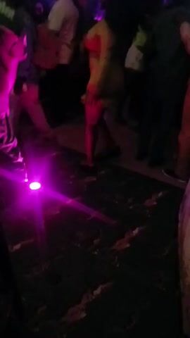 The video shows a crowded, dimly lit club with disco lighting. People are dancing and moving around on a patterned floor. One person is wearing a green shirt with "Parma" on the back. Various colored lights illuminate the scene, including pink, blue, green, and orange. There are other patrons visible, but they are not in clear focus.