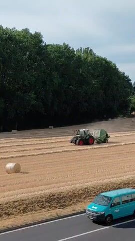 This media file shows an aerial view of a rural landscape in Ecublens, Switzerland. The scene is set in a harvested field of golden wheat, with rows of stubble indicating where the crop was cut. In the middle ground, a green tractor with red accents is actively baling the straw, creating a cylindrical bale of hay and kicking up dust. The tractor is connected to a baler, which is the machine forming the bales. In the foreground, a bright turquoise van with the text "Orelli" is driving on a paved road. Further back, a line of dense green trees forms a backdrop to the field, and hints of buildings and infrastructure can be seen in the distance. The sky is partly cloudy, suggesting it is daytime. The overall impression is of agricultural activity taking place on a sunny day in the Swiss countryside.