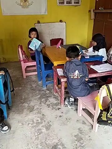 The video shows a classroom with young children and a teacher. The room has yellow walls and a concrete floor. There are wooden tables and chairs, some of which are painted in bright colors. On the walls, there are educational posters and a flag. Children are engaged in various activities, such as drawing or playing. One child is bent over a table, another is sitting at a table with papers, and a third is standing near a table. A female teacher, wearing a headscarf, is sitting at a table and appears to be reading or looking at papers. She is looking towards the camera and seems to be talking. The overall atmosphere appears to be a typical classroom setting for young learners.