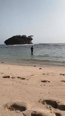The video shows a tranquil beach scene. In the foreground, footprints are visible in the light brown sand. The gentle waves of the ocean wash onto the shore. In the middle ground, a lone person stands in the shallow water, facing away from the camera. In the background, a small, rocky island with sparse vegetation rises from the sea. The sky is clear and bright, suggesting a sunny day. The overall atmosphere is peaceful and serene.