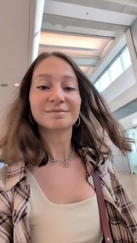 A young woman with shoulder-length brown hair is standing in an indoor setting that appears to be an airport. She is holding up a dark blue passport with Hebrew writing on it in her right hand. She is wearing a light-colored tank top and a plaid shirt over it. She has a necklace with small green beads and a silver pendant. The woman smiles at the camera, then turns her head slightly and speaks to the camera before looking directly into it again. The background shows a bright, modern interior with large windows and ceiling lights.