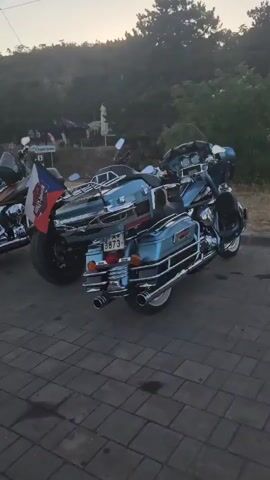 A video shows a line of parked motorcycles, predominantly Harley-Davidsons, on a paved surface. The motorcycles are mostly in a light blue or orange color scheme and feature chrome accents. One motorcycle in the foreground has a Serbian flag attached to its rear. In the background, there are trees and hills, and a few other vehicles and structures are visible, suggesting an outdoor setting like a parking area or roadside stop. The lighting indicates it is either early morning or late afternoon. There are no people clearly visible interacting with the motorcycles.