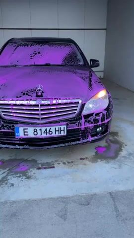 A black Mercedes-Benz sedan is parked inside a car wash bay, completely covered in vibrant purple car wash foam. The foam drips down the sides of the vehicle onto the concrete floor. The car's headlights are illuminated. The Mercedes-Benz star logo is visible on the front grille, and the license plate, which reads "BG E 8146 HH" with the Bulgarian country code and EU flag, is clearly displayed. The setting includes plain walls and a concrete floor, typical of a car wash facility. No individuals are visible in the frame.