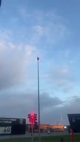 The video shows a seagull perched atop a streetlight. The seagull is white with a light-colored beak, and it stands tall on the lamp fixture. The streetlight is tall and metallic, set against a sky of light blue and scattered clouds. In the background, there are buildings and a traffic light displaying a red "walk" symbol. Signs are visible on the buildings, advertising "Low End Priser" and "Shape Up Fitness." The scene is illuminated by daylight.