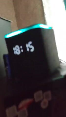 The video shows a close-up of a black digital clock with a glowing cyan light at the top. The clock displays the time "18:15". The background is blurry but appears to be an indoor setting with brown patterned wallpaper and white curtains. The clock is placed on a dark surface, possibly a shelf or table. There is a dark object to the left of the clock, which could be a remote control. The scene is dimly lit, with the primary light source appearing to be the glowing clock.