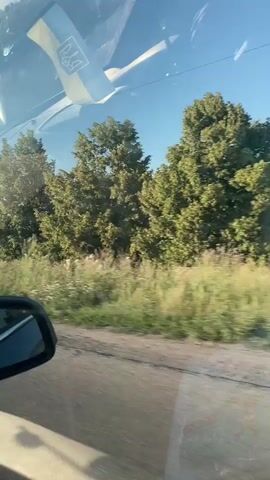 The video shows a view from inside a vehicle, looking out through the windshield and side mirror. A Ukrainian flag with a trident emblem is visible, hanging from the rearview mirror or dashboard. The vehicle appears to be driving on a rural road with a dirt or gravel surface. The landscape outside consists of green fields, trees, and utility poles under a clear blue sky. The sun is shining, casting light and shadows. The perspective suggests the viewer is a passenger in the car, observing the passing scenery.