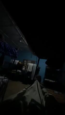 A very dark, grainy video shows what appears to be a dormitory or shared living space at night. The camera is positioned low, close to a bed covered with a patterned blanket or sheet. In the background, bunk beds are visible, with some bedding and what appear to be towels or blankets draped over them. The walls are a light blue color. The lighting is dim, and the image quality is poor, making it difficult to discern specific details. There are no people clearly visible in the frame. The overall impression is one of a humble, perhaps crowded, living environment.