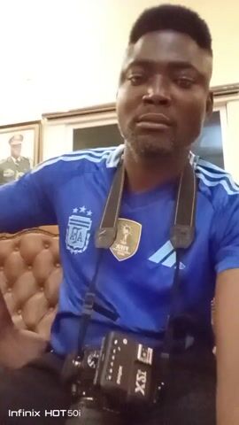 A man wearing a blue soccer jersey with an Argentina logo on the left breast sits on a tufted brown couch, looking into the camera. A camera hangs around his neck. There is a picture on the wall behind him.