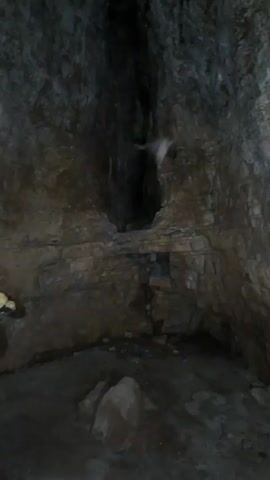 A video shows the interior of a cave with rough, rocky walls and a dark opening in the background. A person's voice is heard stating "Oh, the camera is...". The video then pans to the right, showing more of the cave wall. A second voice is heard saying "Oh, a mouse flew by". The camera continues to pan and a third voice exclaims "Oh, damn, how many?".