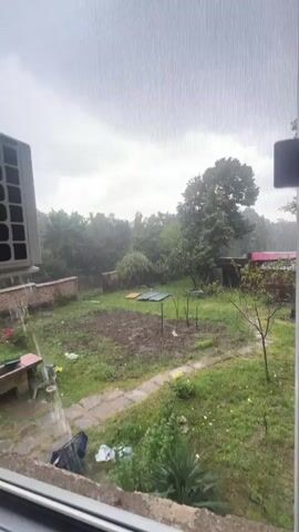 This video shows a backyard scene during a rain shower. The view is from inside, looking through a window screen. In the foreground, to the left, is a part of an air conditioning unit. The backyard features a path, a garden area with tilled soil, some plants, and scattered debris including plastic bags and a blue crate. A brick wall runs along the left side of the yard, and beyond that are trees and foliage. The sky is overcast and gray, indicating the rainy weather. Water is visible in puddles on the ground and is running off surfaces.