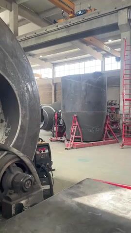 Here's a description of the video:

The video shows a worker welding inside a large metal cylinder. The initial shot is an exterior view of a machine holding the cylinder. The camera moves closer to show the interior. Sparks are visible as the worker welds. The person wears protective gear including a welding helmet, gloves, and a mask. They are crouched inside the cylinder and appear to be working on a seam. The setting appears to be a large industrial workshop.