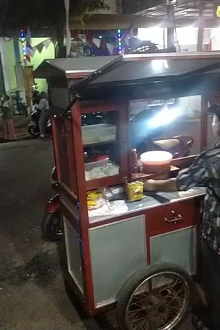 A person wearing a patterned shirt and a dark cap is standing in front of a food cart. The cart is red and has a bicycle wheel on the right side. Inside the cart, there are various items, including containers, packets of food, and what appear to be drinks. The scene is illuminated by artificial lights, suggesting it is nighttime or indoors. In the background, there is a motorcycle and other objects, indicating an outdoor or market setting. Another person is partially visible in the background, near the motorcycle.