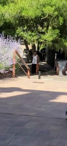 A skateboarder in a dark blue t-shirt, black shorts, and a black baseball cap performs a trick on a skateboard. Another person, wearing a white tank top and brown pants, stands on a skateboard nearby. The scene takes place outdoors on a paved area with some greenery and a railing in the background. The skateboarder in the blue t-shirt approaches a rail and appears to be attempting a grind or a similar trick. The other person is watching. The lighting suggests it is daytime.